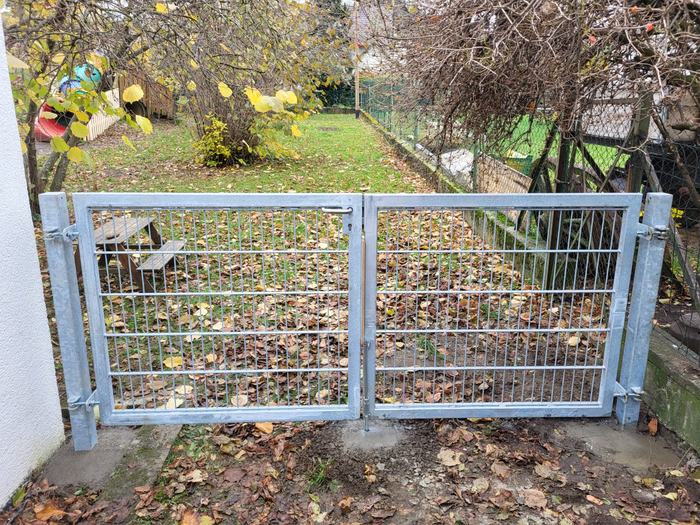 Frontansicht eines zweiflügeligen Metalltores mit Gitterfüllung vor einem Garten.